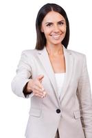 Gefeliciteerd zelfverzekerd jong zakenvrouw in pak uitrekken uit hand- voor beven en glimlachen terwijl staand tegen wit achtergrond foto