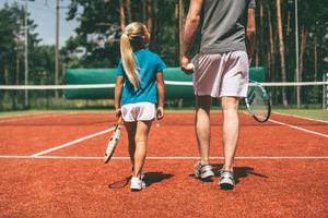 voorbereidingen treffen naar groot spel. achterzijde visie van weinig blond haar- meisje in sport- kleding draag- tennis racket en op zoek Bij haar vader wandelen in de buurt haar door tennis rechtbank foto