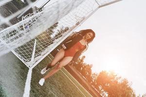 ze liefdes voetbal. vol lengte van aantrekkelijk jong vrouw in rood bikini poseren in de buurt doel post Aan de voetbal veld- foto