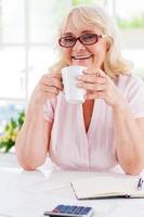 hebben een koffie pauze. gelukkig senior vrouw Holding een kop en glimlachen Bij camera terwijl zittend Bij de tafel foto