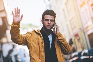 knap jong Mens pratend Aan slim telefoon en golvend naar iemand terwijl staand Aan de stad straat foto