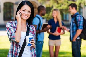 genieten van leerling leven. mooi jong vrouw pratend Aan de mobiel telefoon en glimlachen terwijl staand tegen Universiteit gebouw met haar vrienden chatten in de achtergrond foto