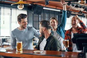 juichen jong mannen in gewoontjes kleding aan het kijken sport spel en genieten van bier terwijl zittend in de kroeg foto