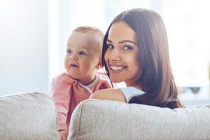 jong mama. achterzijde visie van vrolijk mooi jong vrouw Holding baby meisje in haar handen en op zoek Bij camera met glimlach terwijl zittend Aan de bankstel Bij huis foto