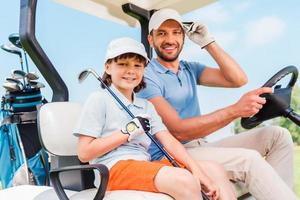 twee glimlachen golfers. gelukkig jong Mens en zijn weinig zoon zittend in golf kar en op zoek Bij camera foto