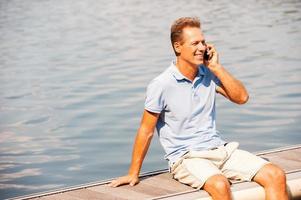 mooi zo praten met vriend. gelukkig volwassen Mens pratend Aan de mobiel telefoon en glimlachen terwijl zittend Bij de kade foto