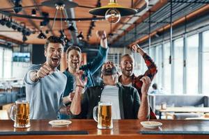 opgewonden jong mannen in gewoontjes kleding drinken bier en aan het kijken sport spel terwijl zittend in de kroeg foto