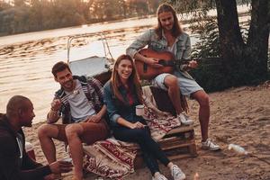 Nee draagt en stormloop. groep van jong mensen in gewoontjes slijtage glimlachen terwijl genieten van strand partij in de buurt de meer foto