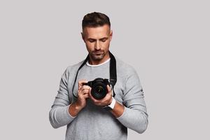 zelfverzekerd fotograaf. mooi zo op zoek jong Mens Holding digitaal camera terwijl staand tegen grijs achtergrond foto