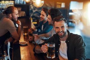 top visie van zorgeloos jong mannen in gewoontjes kleding drinken bier en bonding samen terwijl zittend in de kroeg foto