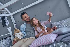 vrolijk vader en zijn weinig dochter nemen selfie terwijl zittend Aan de bed Bij huis foto