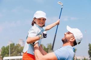 weinig winnaar. vrolijk jong Mens plukken omhoog zijn zoon terwijl staand Aan de golf Cursus foto