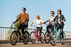 vrienden Aan fietsen. vol lengte van vier jong mensen rijden hun fietsen en glimlachen foto