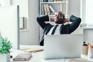 gelukkig jong Afrikaanse Mens in formele kleding glimlachen en houden handen achter hoofd terwijl werken in de kantoor foto