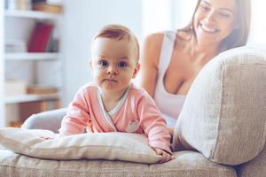 schattig met mama. weinig baby meisje op zoek Bij camera en leunend Aan kussen terwijl haar moeder zittend Aan de bankstel Bij achtergrond foto