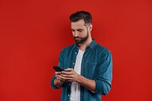 knap jong Mens in gewoontjes kleding gebruik makend van slim telefoon en glimlachen terwijl staand tegen rood achtergrond foto