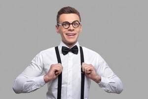 zelfverzekerd nerd. portret van jong nerd Mens in boog stropdas aanpassen zijn bretels en glimlachen terwijl staand tegen grijs achtergrond foto