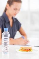 Nee tijd voor lunch. mooi jong vrouw schrijven iets in haar Notitie stootkussen terwijl belegd broodje en een fles van water houdende Aan voorgrond foto