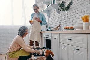 gelukkig senior paar in schorten voorbereidingen treffen gezond avondeten in oven en glimlachen terwijl uitgeven tijd Bij huis foto