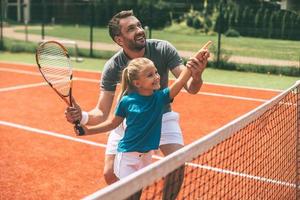 tennis is pret wanneer vader is in de buurt. vrolijk vader in sport- kleding onderwijs zijn dochter naar Speel tennis terwijl beide staand Aan tennis rechtbank foto