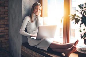 schattig Bij huis. mooi jong vrouw gebruik makend van haar laptop en op zoek Bij camera met glimlach terwijl zittend Bij vensterbank Bij huis foto