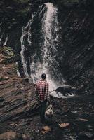 iedereen zou moeten zien het. vol lengte achterzijde visie van jong Mens op zoek Bij de waterval terwijl wandelen in de bergen foto