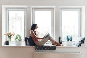 zorgeloos jong vrouw in knus pyjama lezing een boek terwijl resting Aan de venster dorpel Bij huis foto