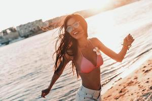 extreem sexy. aantrekkelijk jong vrouw in bikini glimlachen en op zoek weg terwijl wandelen Aan de strand buitenshuis foto