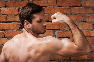 tonen zijn perfect biceps. achterzijde visie van zelfverzekerd jong gespierd Mens poseren terwijl staand tegen steen muur foto