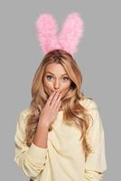 oeps... aantrekkelijk jong vrouw in roze konijn oren aan het bedekken mond met hand- en op zoek Bij camera terwijl staand tegen grijs achtergrond foto