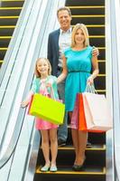 familie Aan boodschappen doen Spree. vrolijk familie Holding boodschappen doen Tassen en glimlachen Bij camera terwijl in beweging door roltrap foto