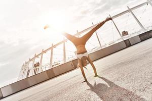 fantastisch gymnastiek. vol lengte van modern jong vrouw in sport- kleding aan het doen handstand terwijl oefenen buitenshuis foto