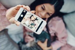 top visie van mooi jong vrouw in pyjama omarmen huiselijk kat en nemen selfie terwijl resting in bed Bij huis foto