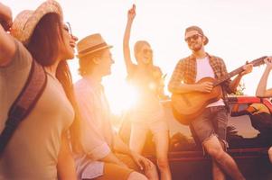 weg reis is plezier. groep van jong vrolijk mensen dansen en spelen gitaar terwijl genieten van hun weg reis in oppakken vrachtauto samen foto