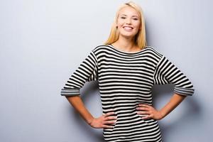 glimlachen vertrouwen. mooi jong blond haar- Dames Holding handen Aan heup en op zoek Bij camera met glimlach terwijl staand tegen grijs achtergrond foto