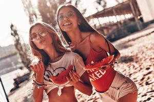 lui heet dag. twee aantrekkelijk jong Dames glimlachen en aan het eten watermeloen terwijl staand Aan de strand foto
