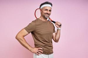 vrolijk volwassen Mens draag- tennis racket Aan schouder terwijl staand tegen roze achtergrond foto