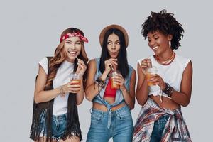 drie aantrekkelijk jong Dames drinken verfrissend cocktail en glimlachen terwijl staand tegen grijs achtergrond foto