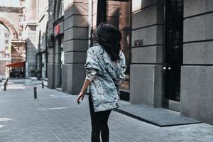 stad stijl. achterzijde visie van jong vrouw in denim jasje wandelen naar beneden de straat foto