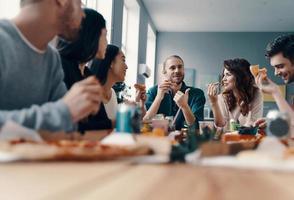 leven is beter met vrienden. groep van jong mensen in gewoontjes slijtage aan het eten pizza en glimlachen terwijl hebben een avondeten partij binnenshuis foto