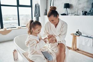 moeder met dochter in badjassen glimlachen en gebruik makend van hand- room terwijl aan het doen ochtend- routine- foto