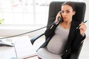 zwanger bedrijf dame. mooi zwanger zakenvrouw pratend Aan de telefoon terwijl zittend Bij haar werken plaats in kantoor foto