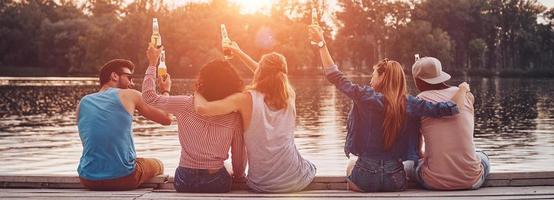 achterzijde visie van jong mensen in gewoontjes slijtage roosteren met een bier flessen terwijl zittend Aan de pier foto