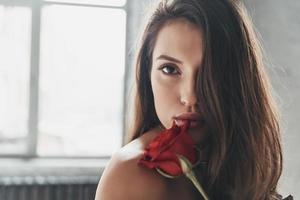 prachtig schoonheid. aantrekkelijk jong vrouw Holding een rood roos en op zoek Bij camera terwijl uitgeven tijd Bij huis foto