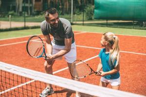 beoefenen tennis. vrolijk vader in sport- kleding onderwijs zijn dochter naar Speel tennis terwijl beide staand Aan tennis rechtbank foto