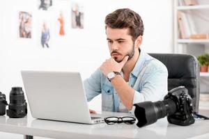 genereren nieuw ideeën. geconcentreerd jong Mens Holding hand- Aan kin en op zoek Bij laptop terwijl zittend Bij zijn werken plaats foto