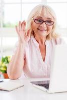 gezegde Hoi naar haar dichtstbijzijnde. vrolijk senior vrouw golvend met haar hand- terwijl op zoek Bij laptop foto