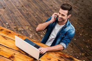 bedrijf Aan vers lucht. top visie van gelukkig jong Mens werken Aan laptop en pratend Aan de mobiel telefoneren zittend Bij de houten tafel buitenshuis foto