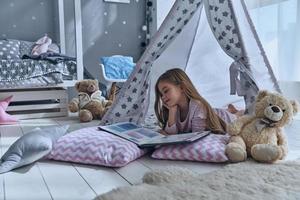 verloren in haar sprookje. schattig weinig meisje lezing boek terwijl aan het liegen Aan de verdieping in tent foto