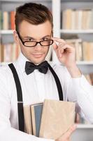 ik weten ik ben de slimste. zelfverzekerd jong Mens in overhemd en boog stropdas Holding boeken en aanpassen bril terwijl staand in bibliotheek foto